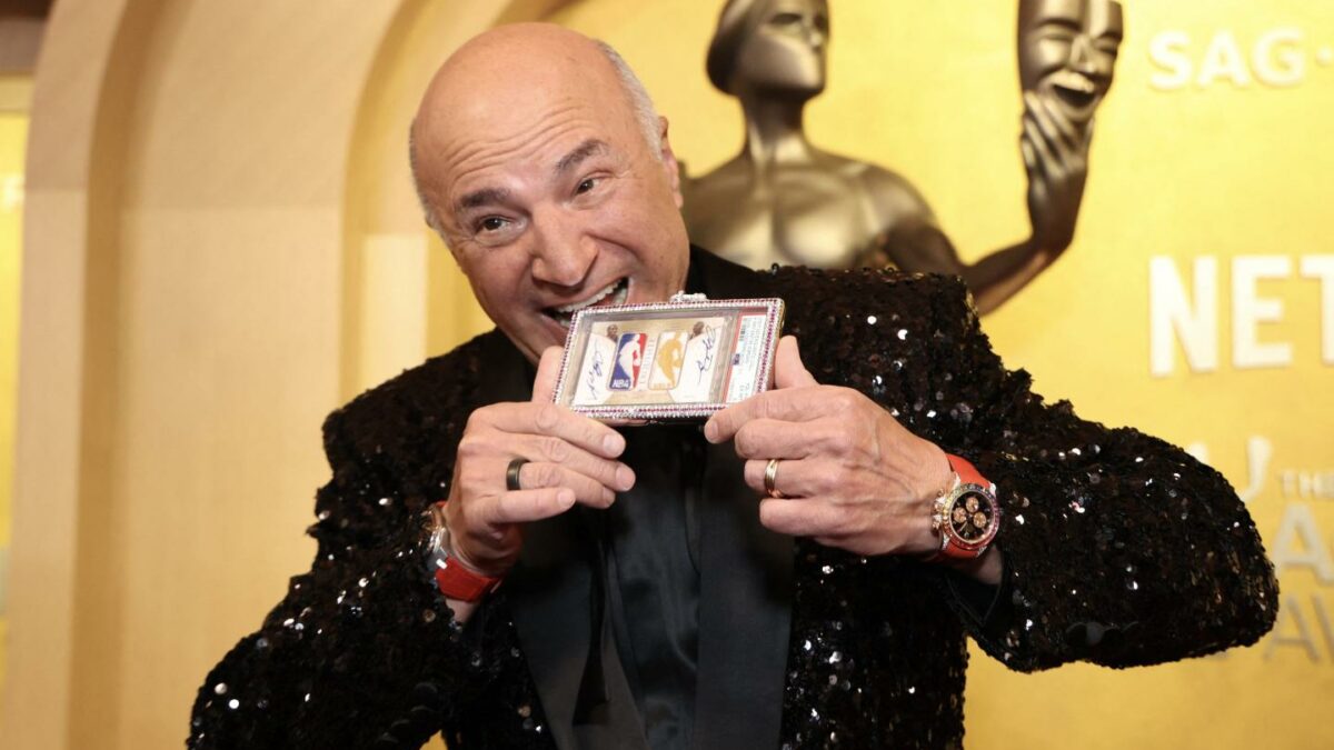 Kevin O'Leary wearing a black suit with a gold NBA trading card necklace in his mouth